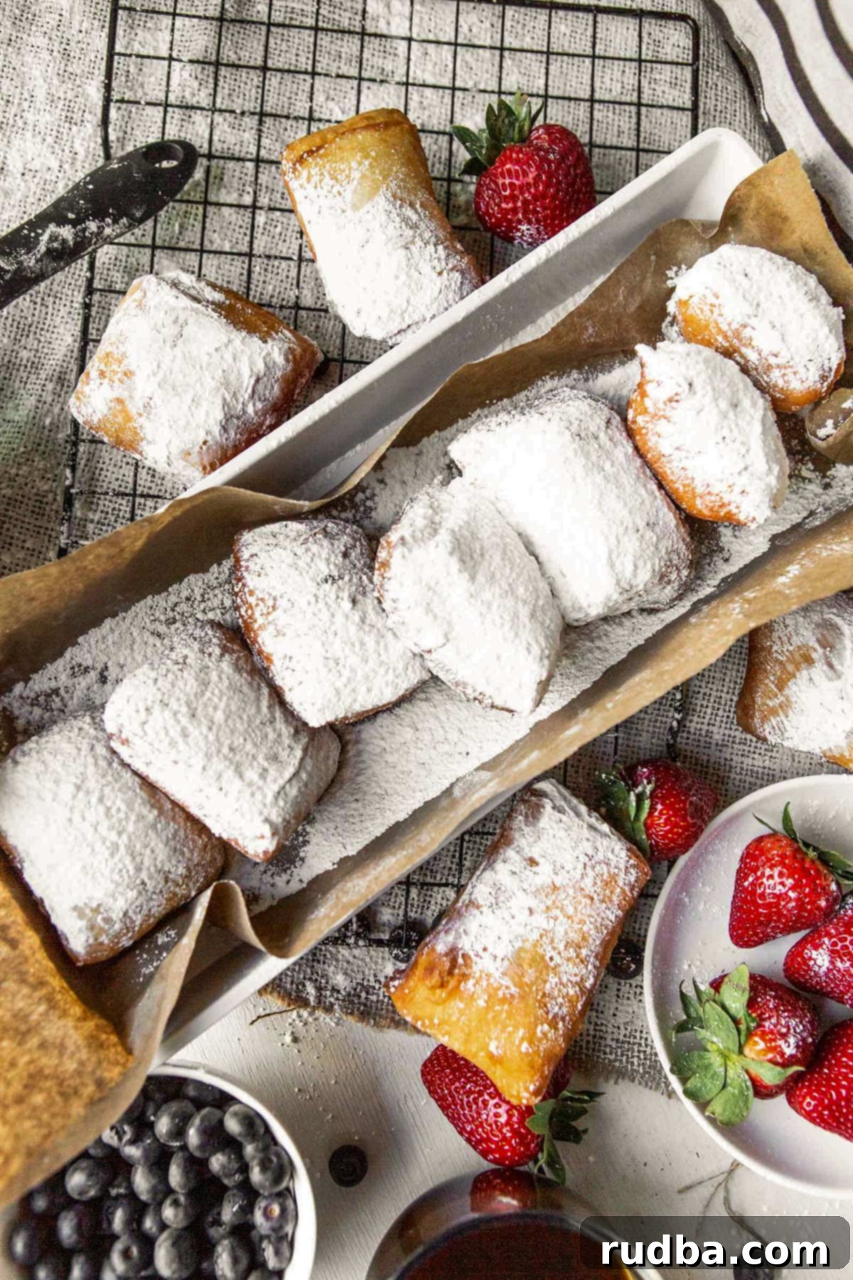 a long bog with brown paper and beignets inside. beignets on the side of the box along with strawberries and blueberries.