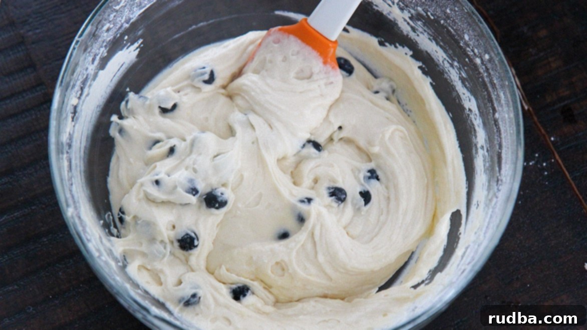 Cake batter being folded with fresh blueberries using a spatula, showing the rich texture before baking.