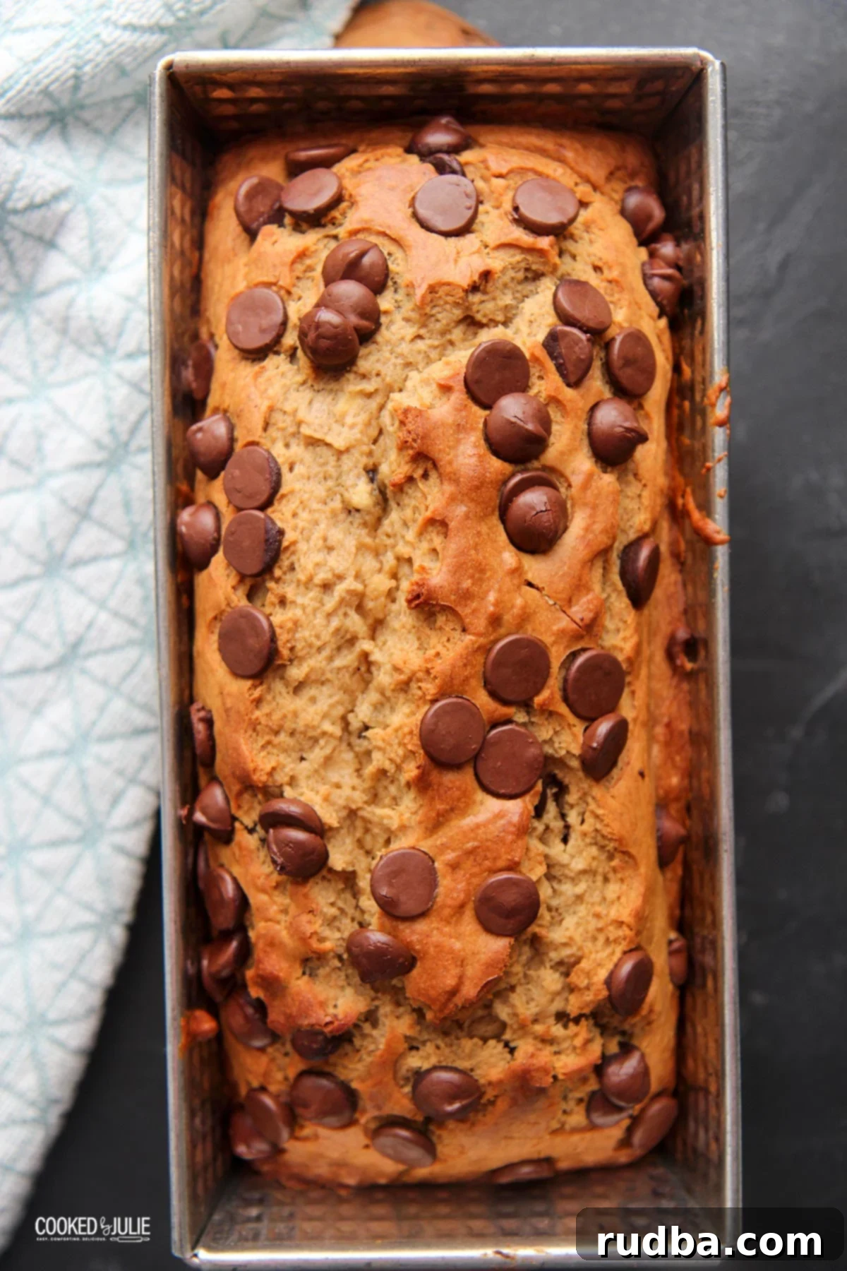 Delicious chocolate chip banana bread in a loaf pan, perfectly golden brown, sitting on a dark surface with a blue linen towel beside it. 