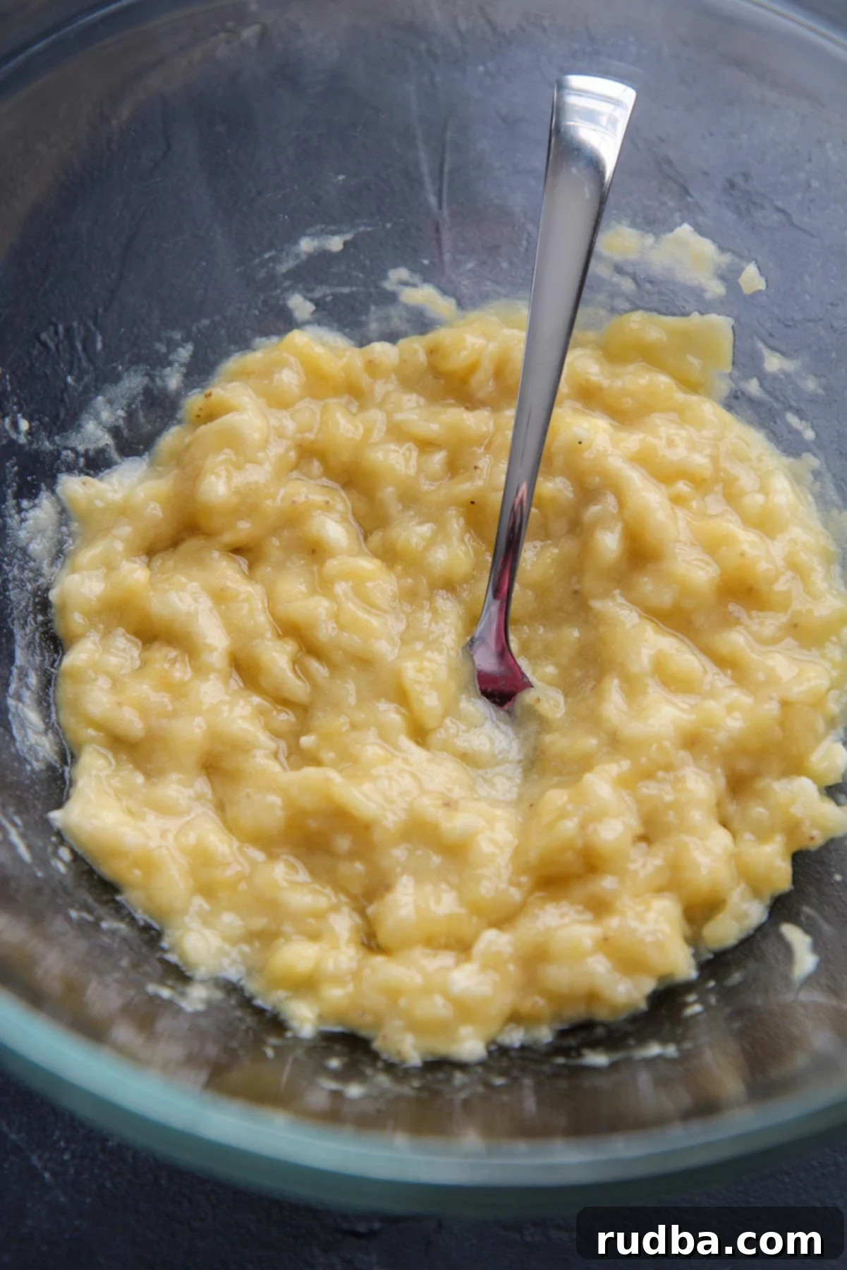 Four perfectly ripe bananas mashed in a clear glass bowl with a fork, ready for baking on a dark kitchen counter.