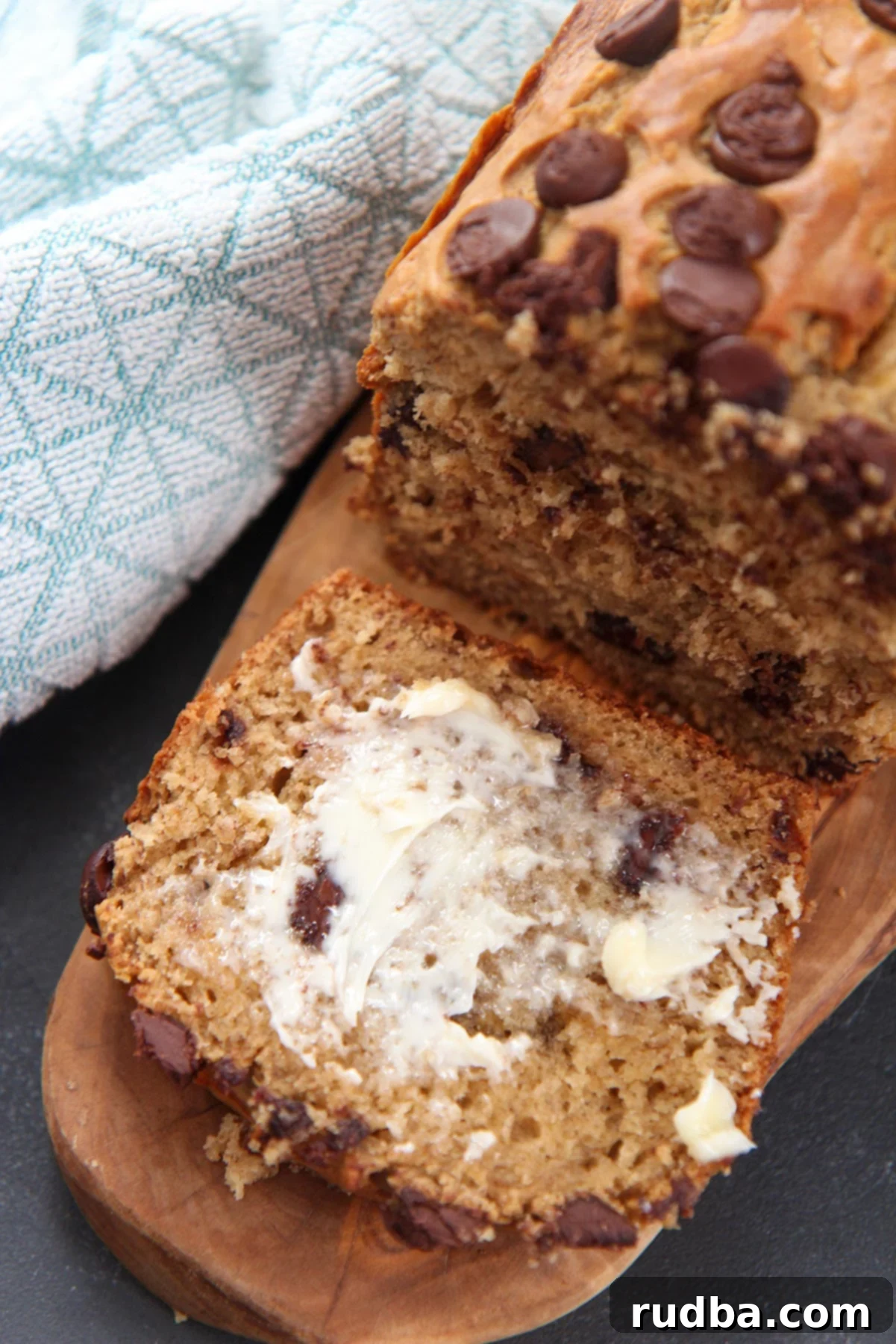 A delectable slice of warm chocolate chip banana bread, butter generously smeared on top, with a blue towel nearby.