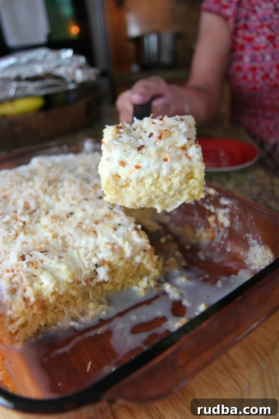 a hand lifting a slice of coconut tres leches and a full cake in the background. 
