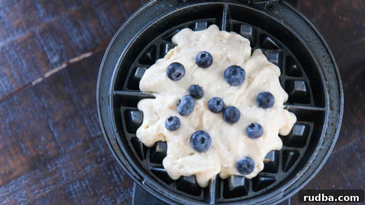 Close-up shot of waffle batter being poured into a waffle iron, with fresh blueberries scattered on top, ready for cooking.