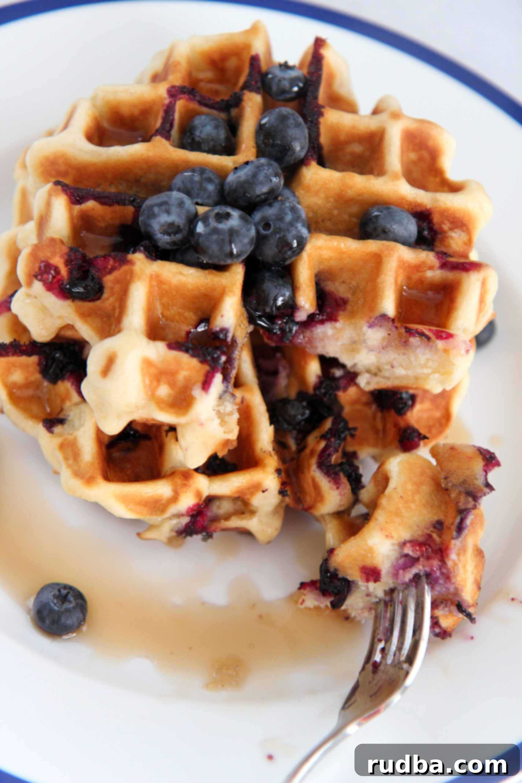 Two stacked buttermilk blueberry waffles on a white plate, with a fork gently slicing into the top waffle, revealing its fluffy interior and blueberries.
