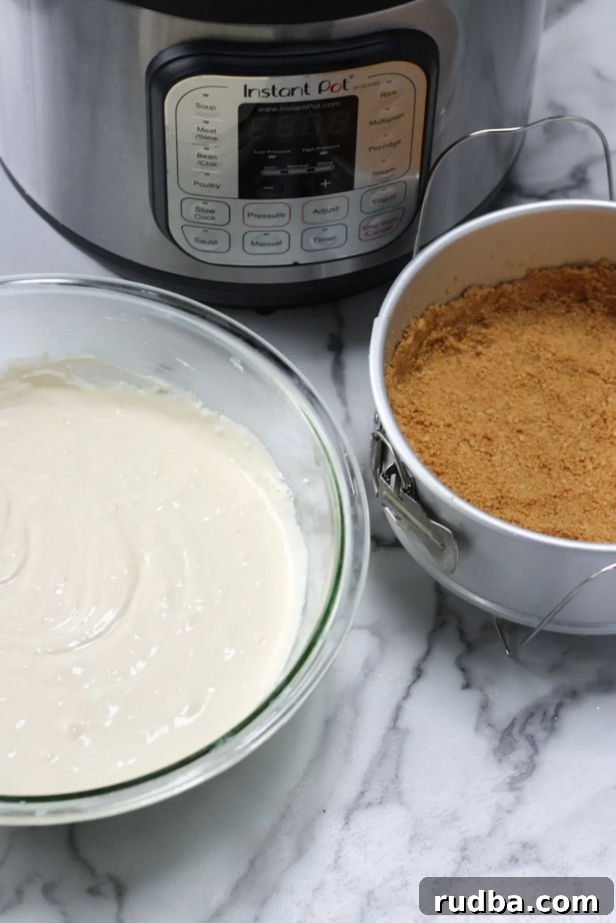 Cheesecake batter in a glass bowl, a springform pan with a graham cracker crust, and an Instant Pot in the background, illustrating the preparation process.
