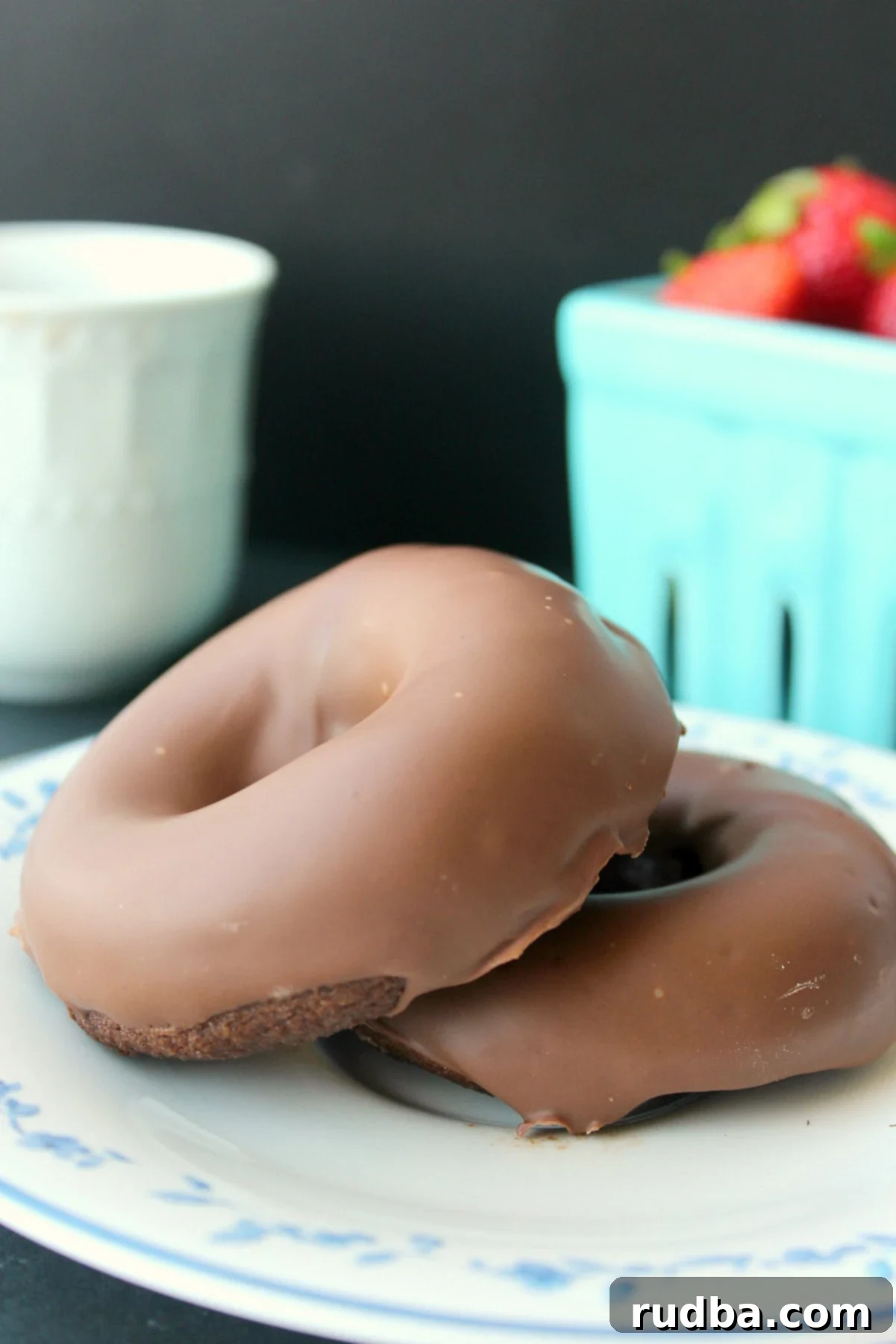 two keto baked chocolate donuts on a white plate with a basket of strawberries in the background. 