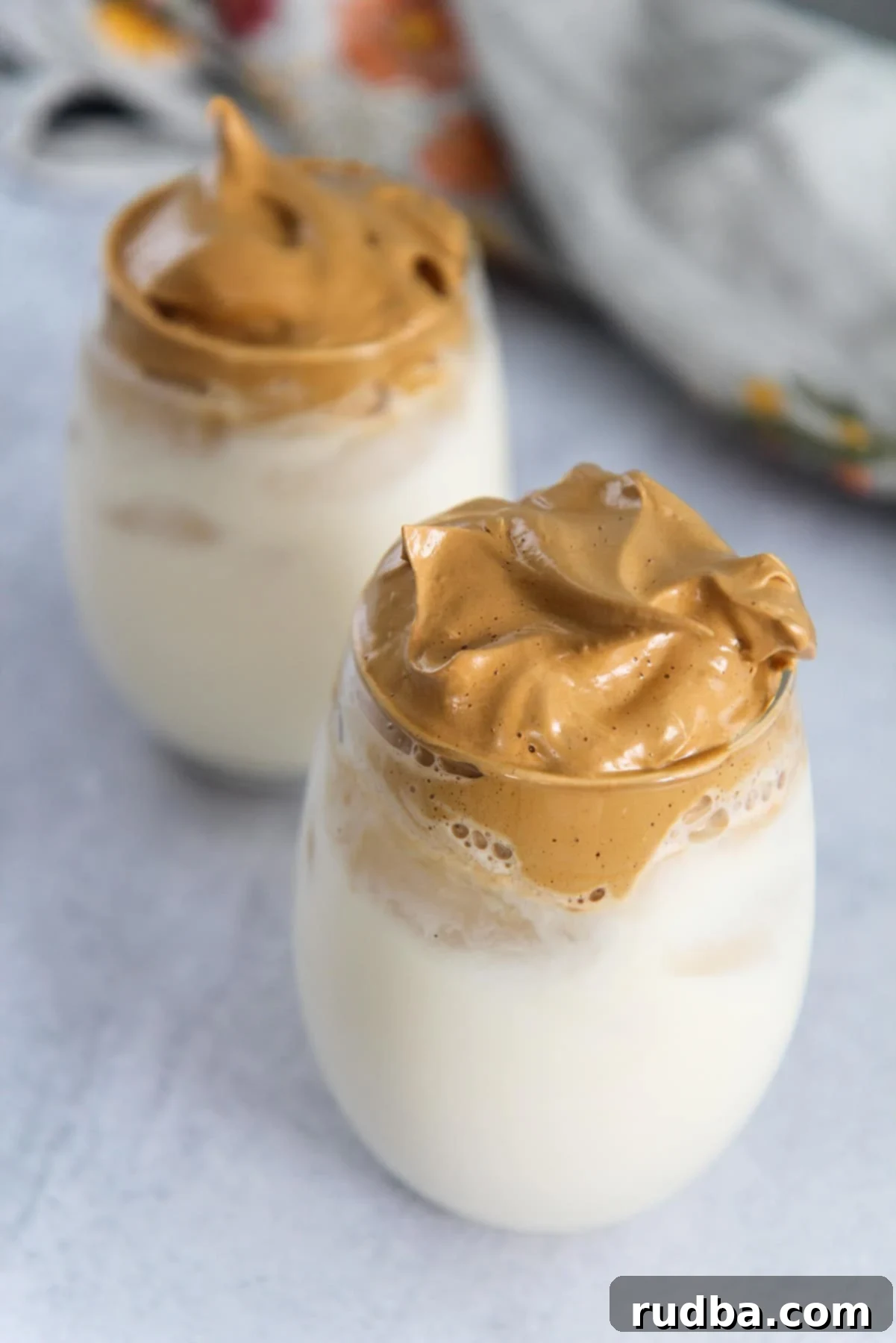Two glasses of whipped coffee on a gray surface with a gray towel in the background, showing the distinct layers of whipped foam and milk.