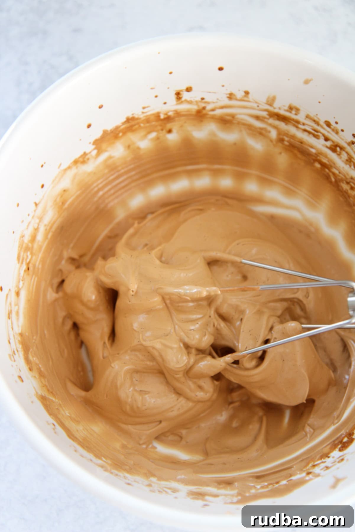 Light brown whipped coffee in a white bowl with a metal whisk, showing its thick, creamy consistency.