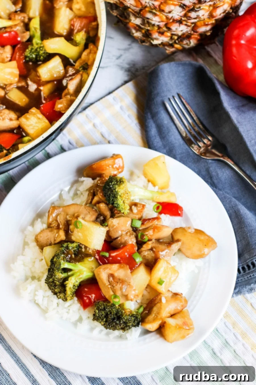Delicious pineapple chicken stir-fry served over fluffy white rice on a white plate, with a fork and a skillet in the background, showcasing a perfect weeknight meal.
