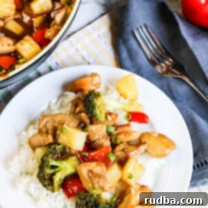 pineapple chicken stir-fry over white rice on a white plate with a fork on the side and skillet in the background.