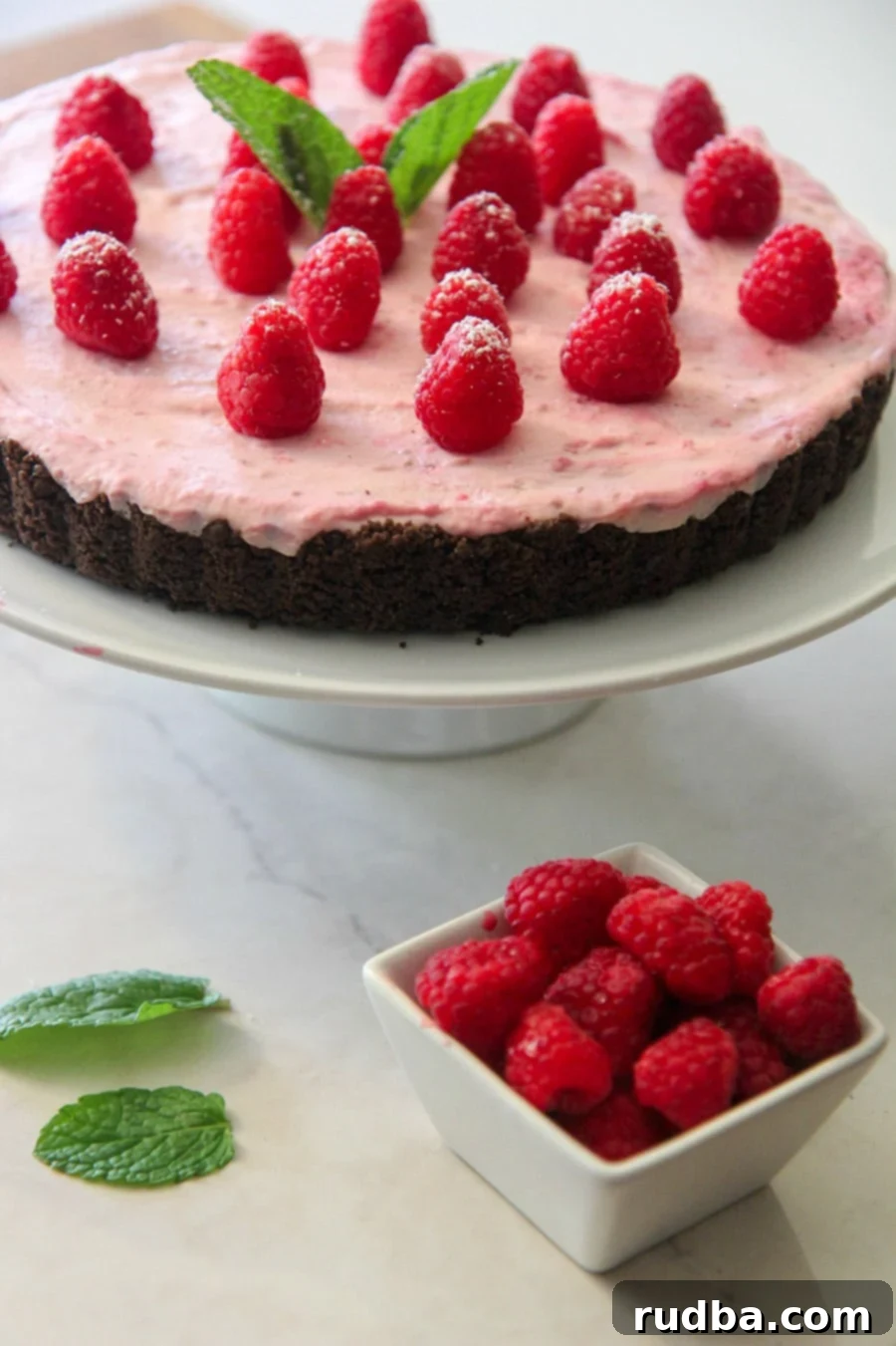 A beautifully presented no-bake white chocolate raspberry tart on a white serving platter, garnished with fresh raspberries and a sprig of mint, ready to be enjoyed.