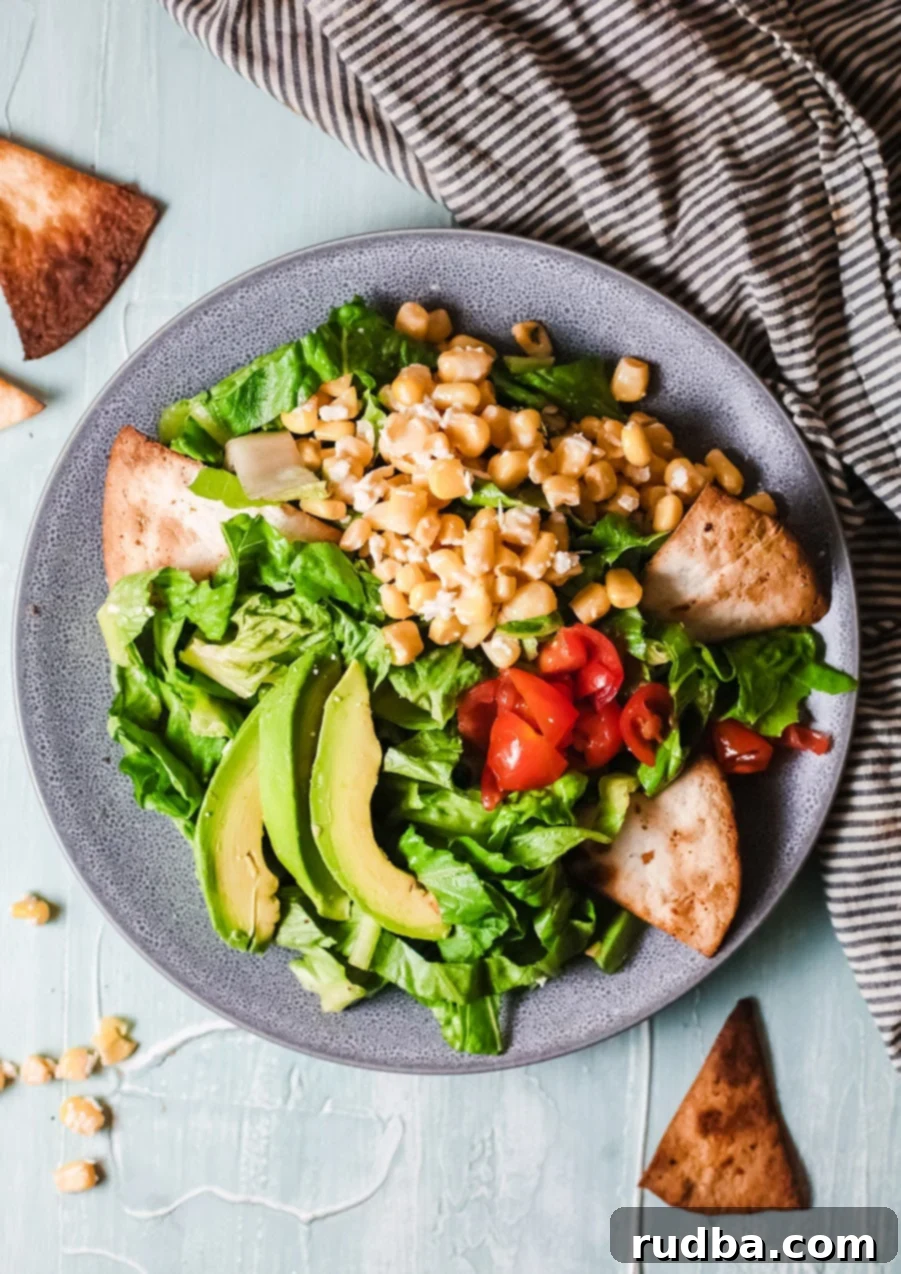 A blue bowl filled with fresh lettuce, vibrant corn kernels, sliced avocado, and colorful tomatoes, with a side of crispy tortilla chips, all placed on a table with a black and white striped towel.