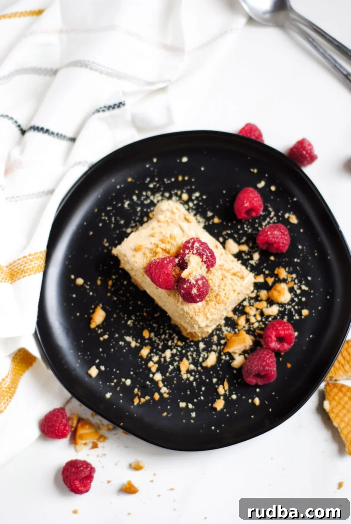 A perfectly squared slice of no-bake Dalgona cheesecake on a sleek black plate, garnished with fresh raspberries and a sprinkle of crushed cookies. A white and yellow striped towel rests casually to the side, completing the inviting scene.