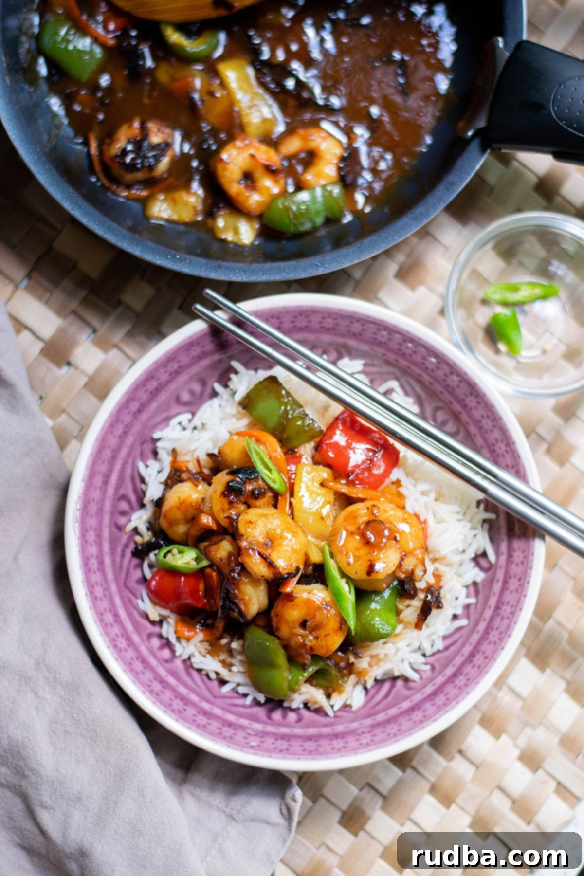 shrimp and rice with chop sticks in a purple bowl with a pan in the background with shrimp, veggies, and sauce.
