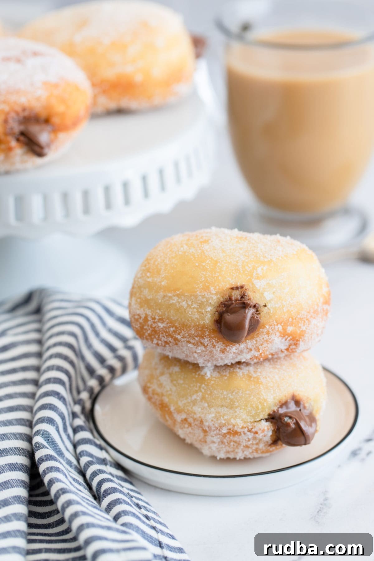 two nutella stuffed donuts on a small white plate with a blue and white towel on the side, a coffee and more donuts in the background.