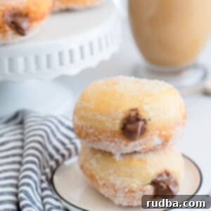 Decadent Nutella-Filled Donuts 5 two nutella stuffed donuts on a small white plate with a blue and white towel on the side, a coffee and more donuts in the background.