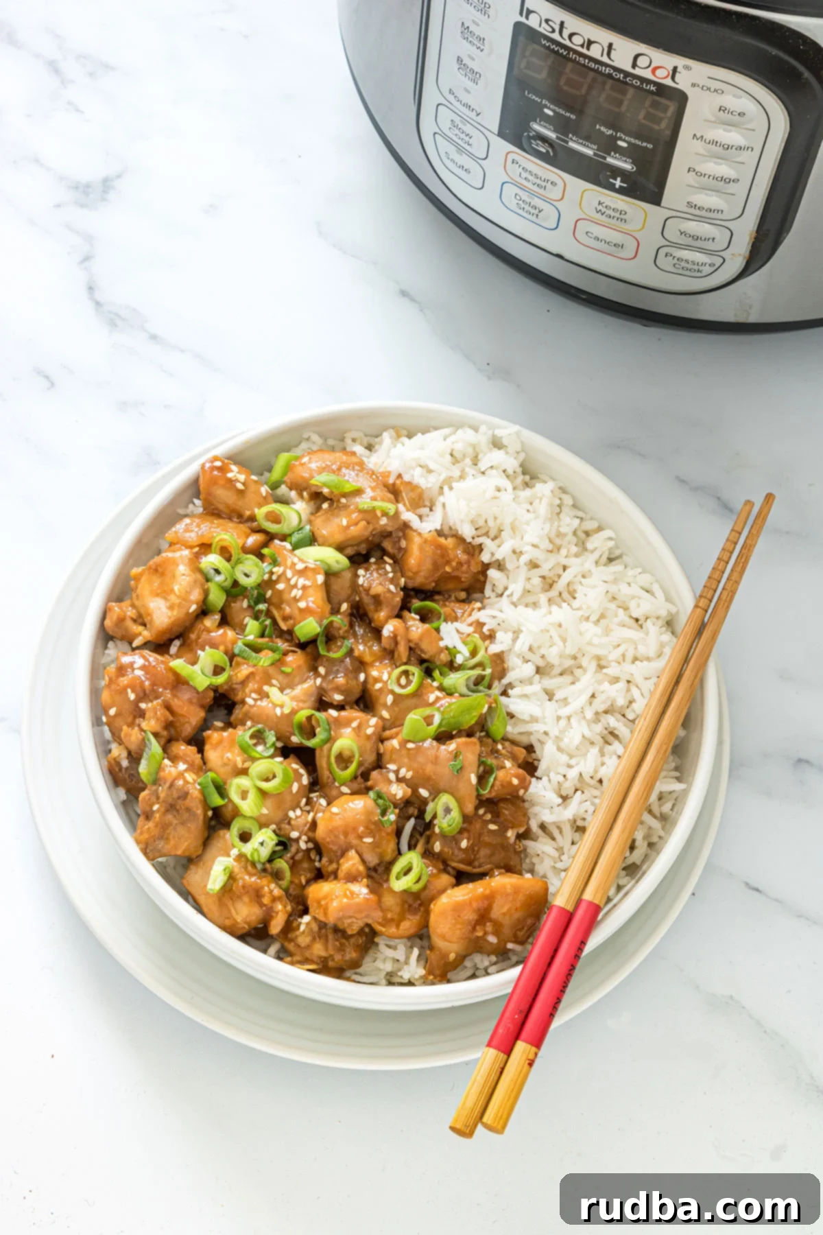 sesame chicken with a side of white rice in a white bowl with chop sticks on top and an instant pot in the background. 