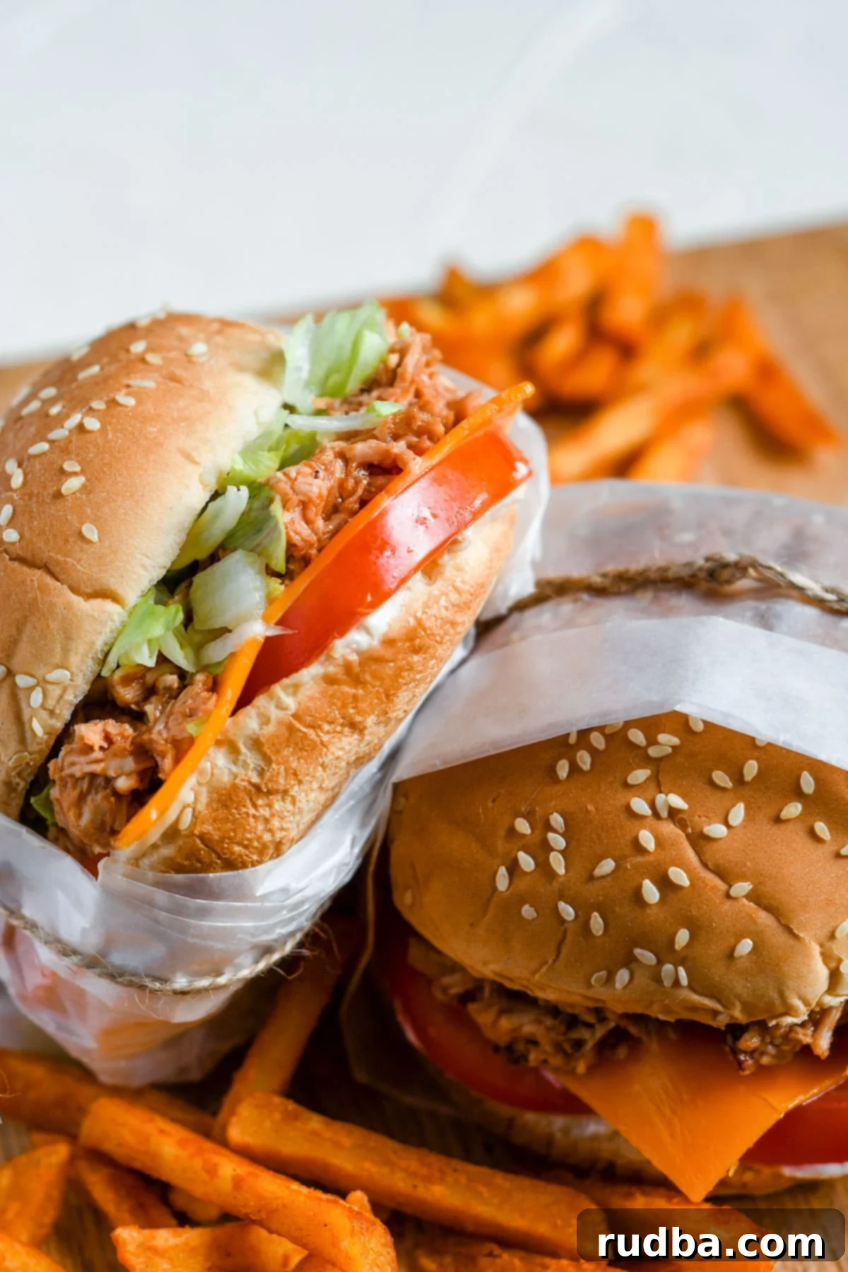 Two mouth-watering shredded chicken sandwiches, generously loaded with lettuce, tomato, and melted cheddar cheese, served alongside a pile of crispy sweet potato fries on a wooden board.