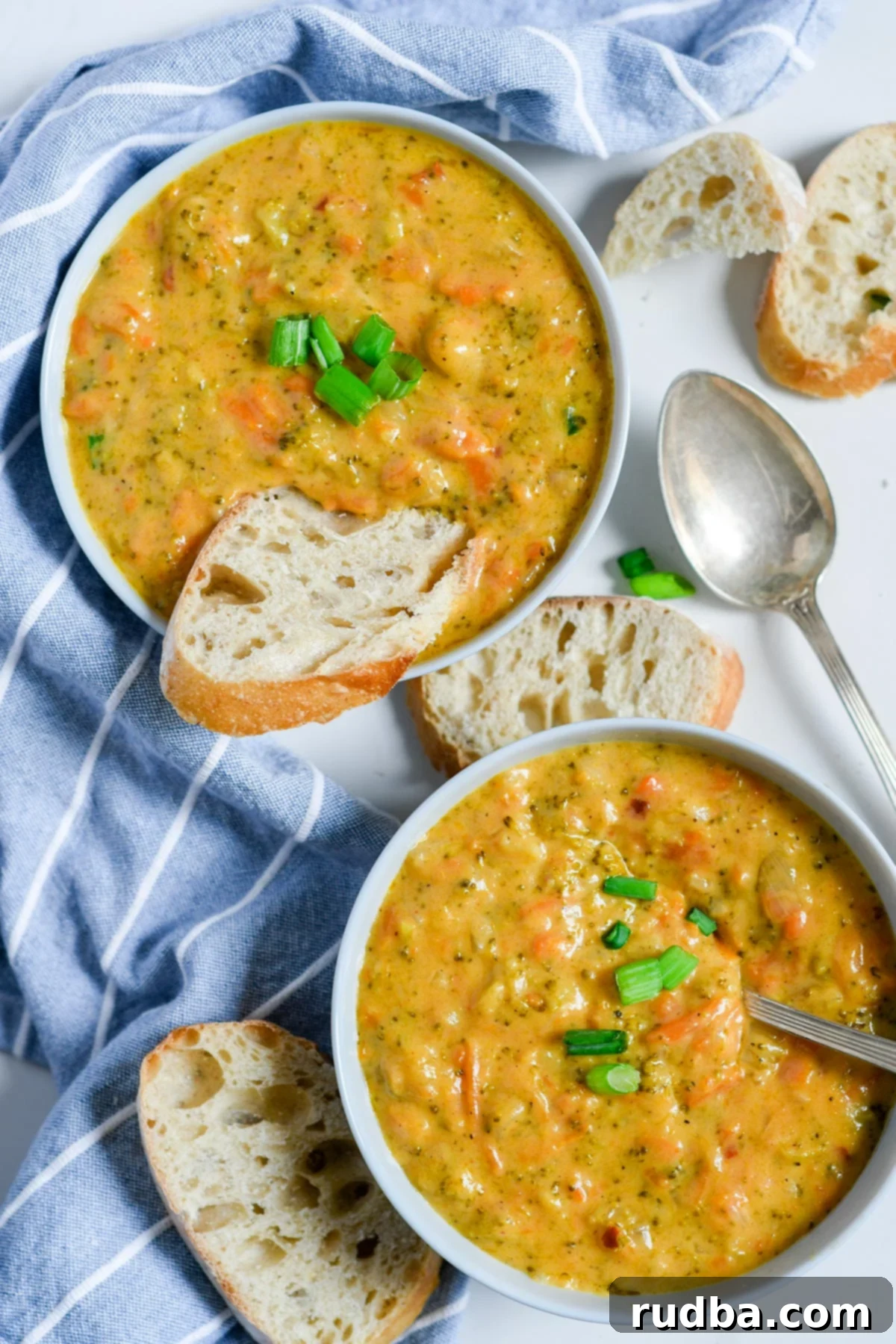 two white bowls with broccoli cheddar soup, slices of bread inside one bowl and on the side. A spoon on the side and a blue and white towel. 