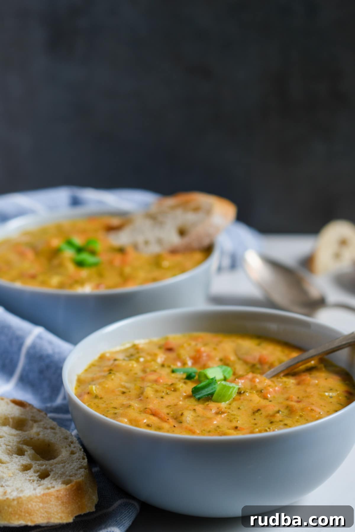two bowls filled with broccoli and cheese soup with scallions on top, bread on the side, a blue and white towel, and a dark backdrop. 