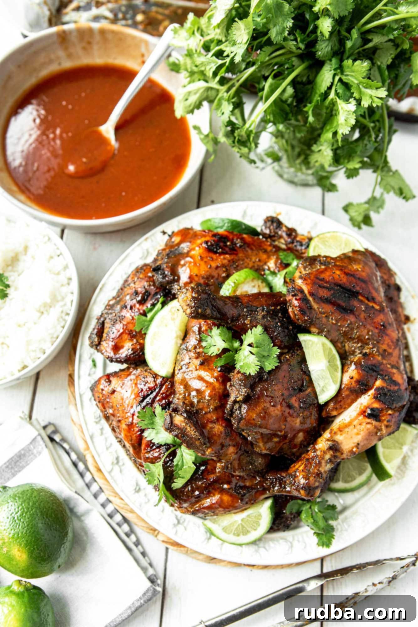 Jamaican jerk chicken on a white plate with cilantro and lime wedges. A bowl of red sauce with a spoon and more cilantro and lime on the side. 
