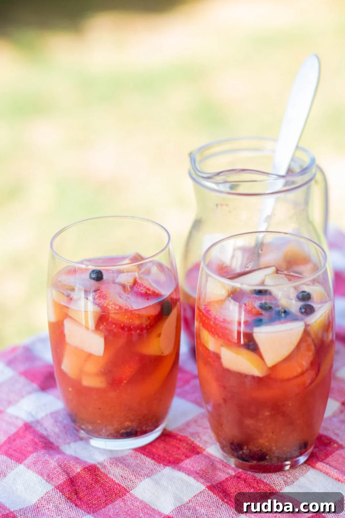 two glasses filled with sangria and fruit on top of a red and white table cloth and a pitcher with sangria in the background. 