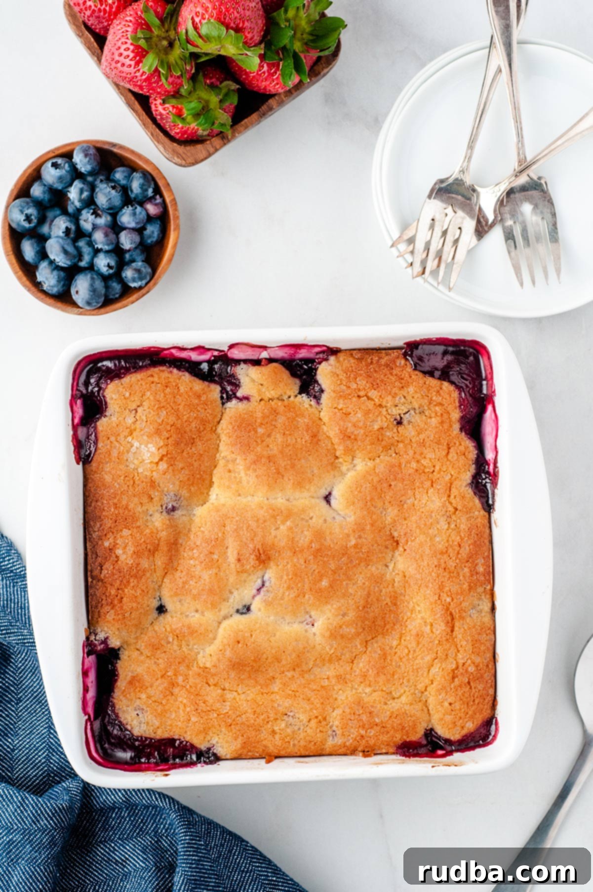 A beautifully baked berry cobbler in a white baking dish, garnished with fresh strawberries and blueberries on the side. Forks and a small white plate are elegantly arranged next to it, inviting a taste.