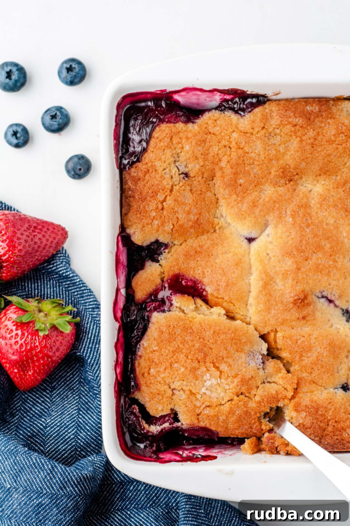 A close-up shot of half a white baking dish filled with a golden-brown, baked berry cobbler. Fresh blueberries, strawberries, and a blue kitchen towel are artfully arranged alongside the dish, highlighting the dessert's freshness and appeal.