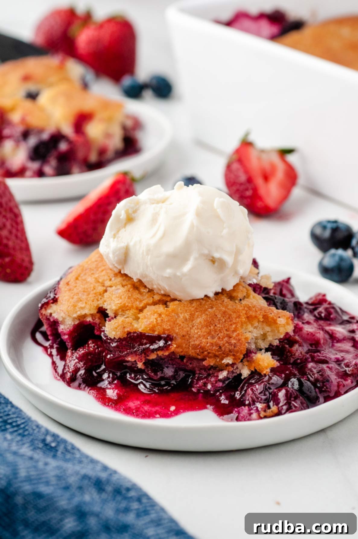 A close-up shot of a single serving of berry cobbler on a white plate, topped with a generous scoop of melting vanilla ice cream. In the softly blurred background, a larger baking dish of cobbler and scattered fresh berries hint at the bounty.