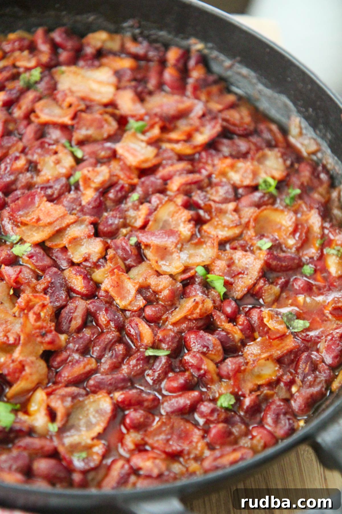 Chipotle baked beans in an iron skillet with crispy bacon and fresh parsley on top. A close-up view highlighting the rich sauce and textures.
