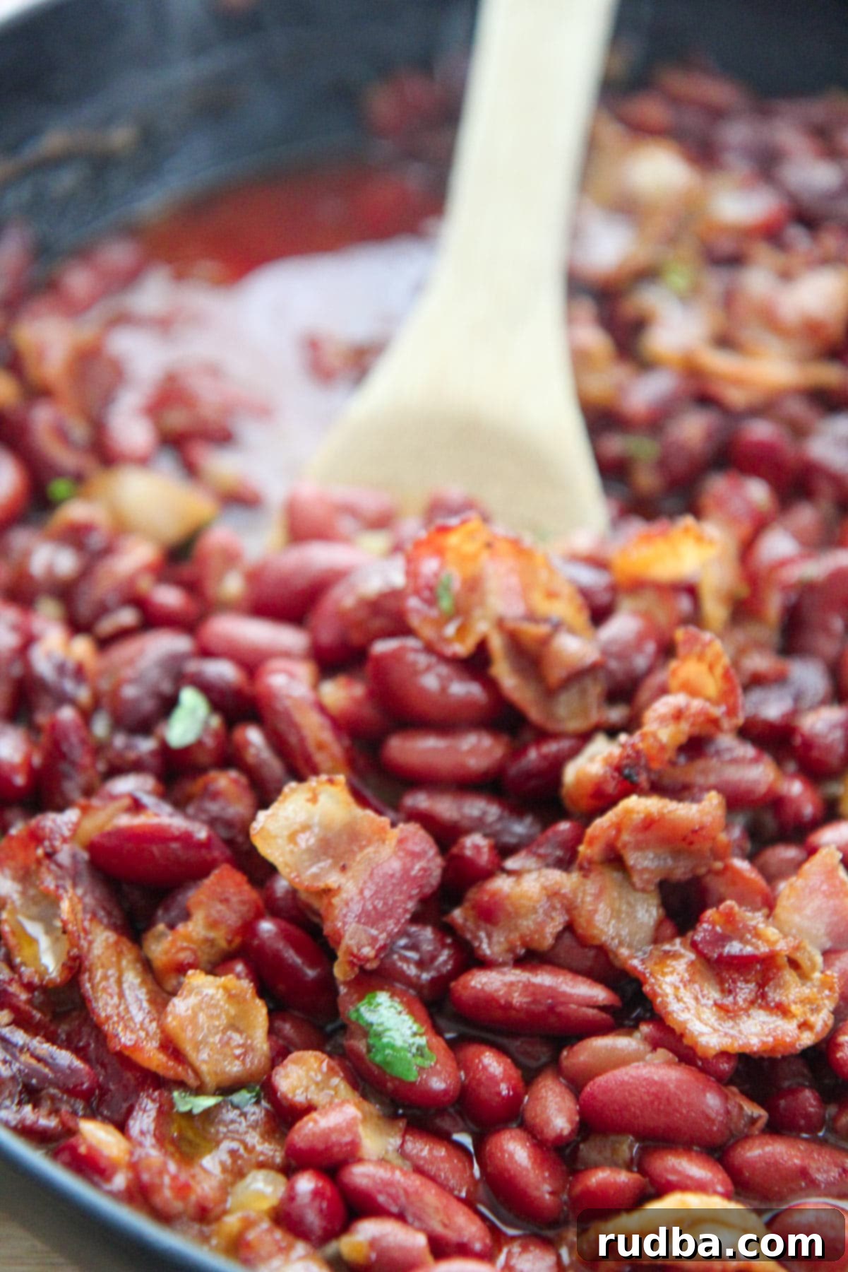 Baked beans in a cast iron skillet with a wooden spoon, ready to be served. A rustic and inviting presentation.