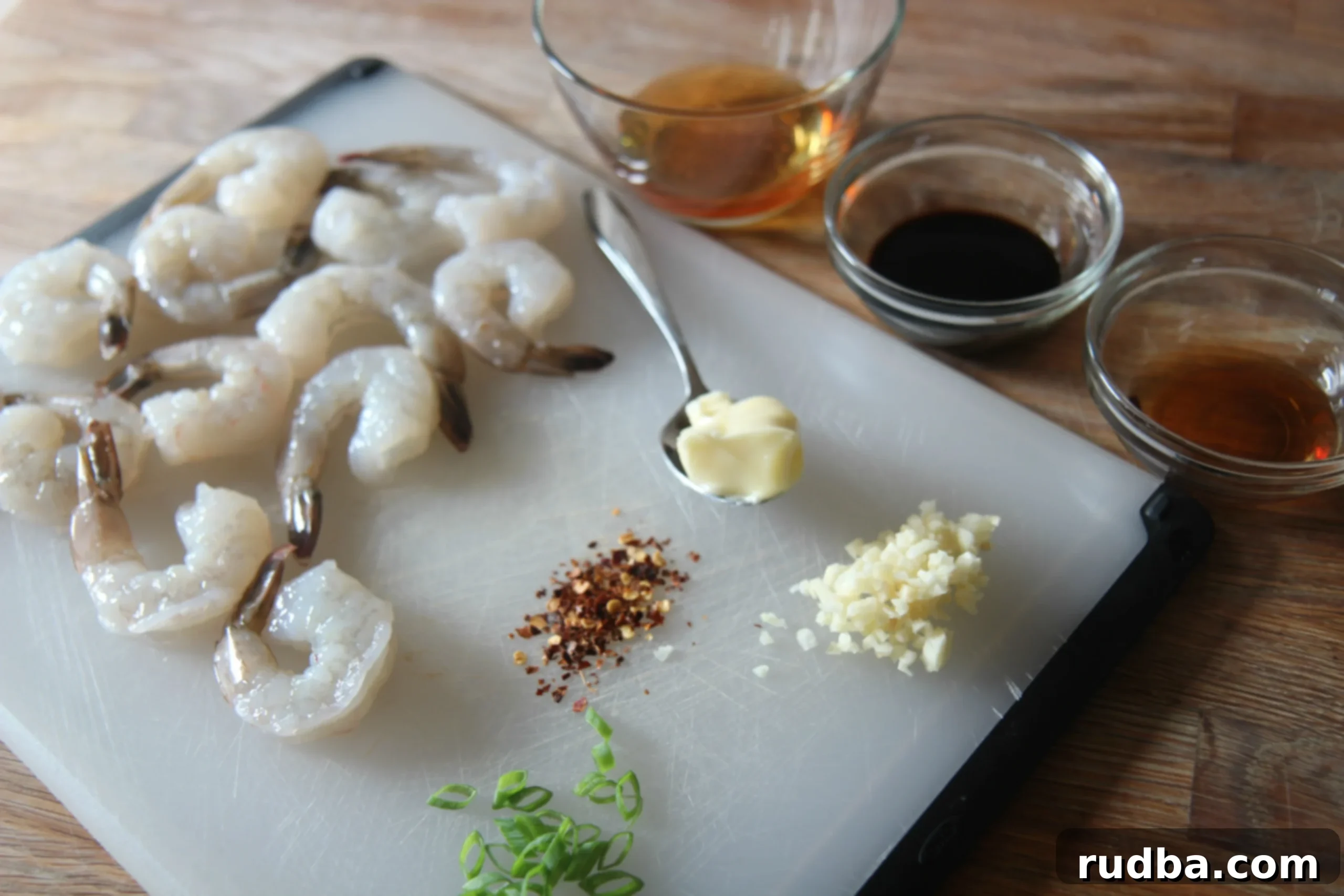 A selection of raw ingredients for honey garlic shrimp arranged on a wooden chopping board: raw shrimp, softened butter, minced garlic, various sauces, and chopped scallions.