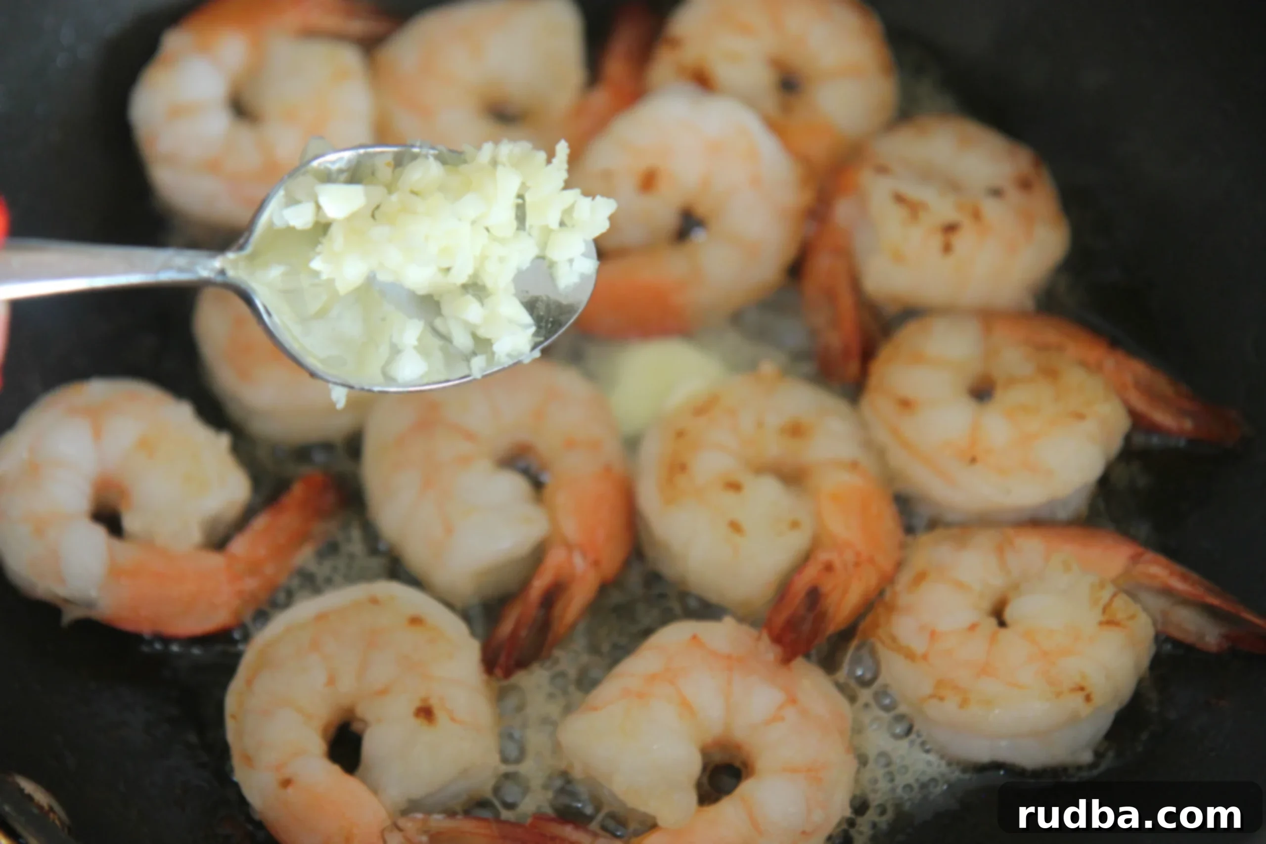 A close-up shot of a spoon of fresh minced garlic being added to shrimp cooking in a skillet.