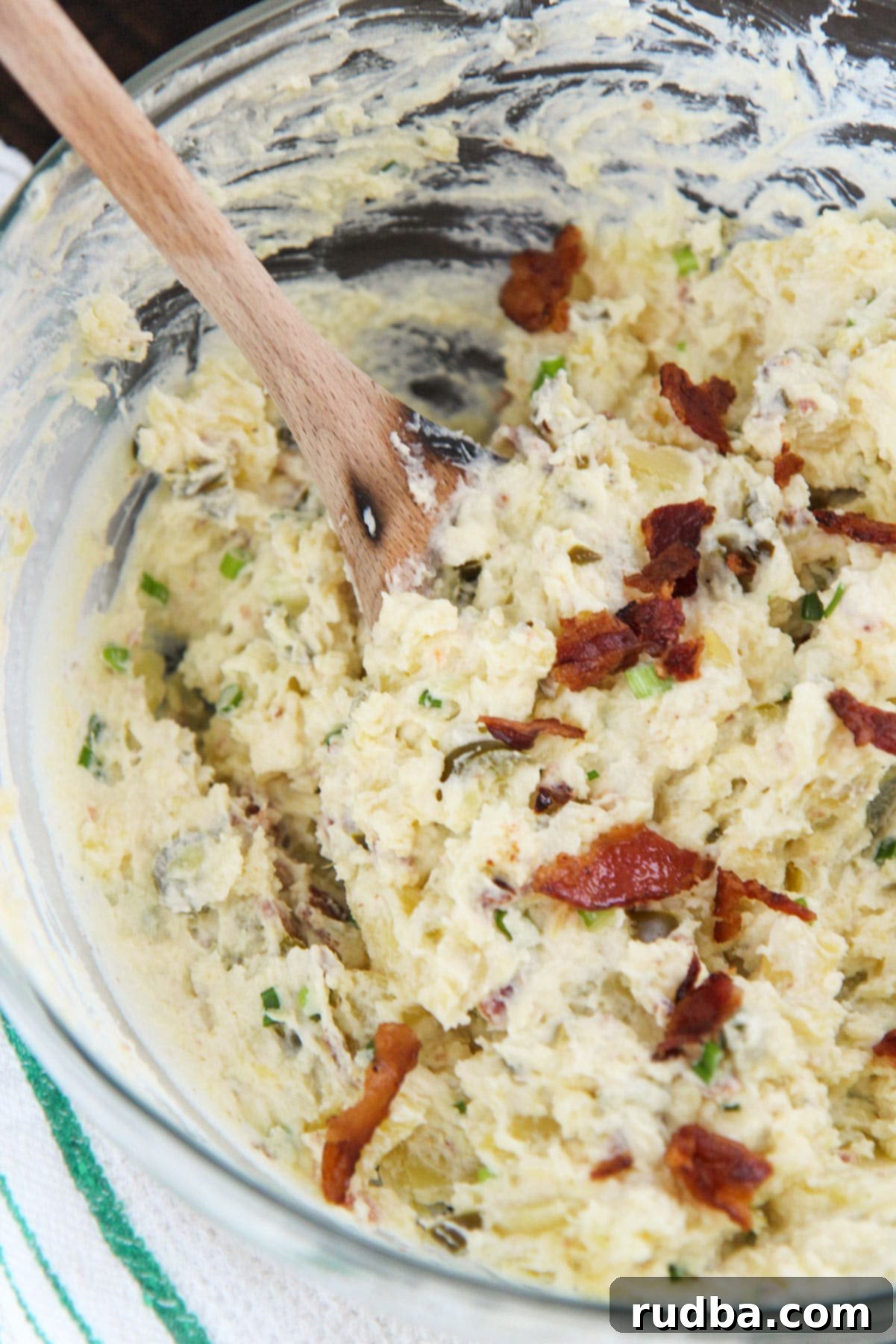 A vibrant spicy potato salad in a clear glass bowl, garnished with extra crispy bacon bits on top. A rustic wooden spoon rests inside the bowl, and a green and white striped towel is neatly folded on the side, hinting at a fresh, home-cooked meal.