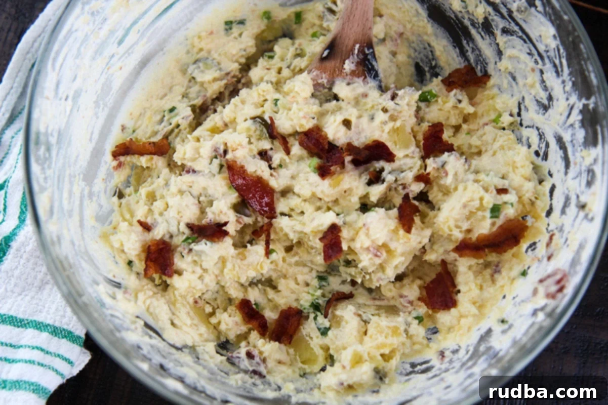 A close-up shot of the creamy spicy potato salad in a glass bowl, showcasing the tender potatoes, visible bits of bacon, and the rich dressing. A patterned towel is seen to the side, adding to the homely aesthetic.