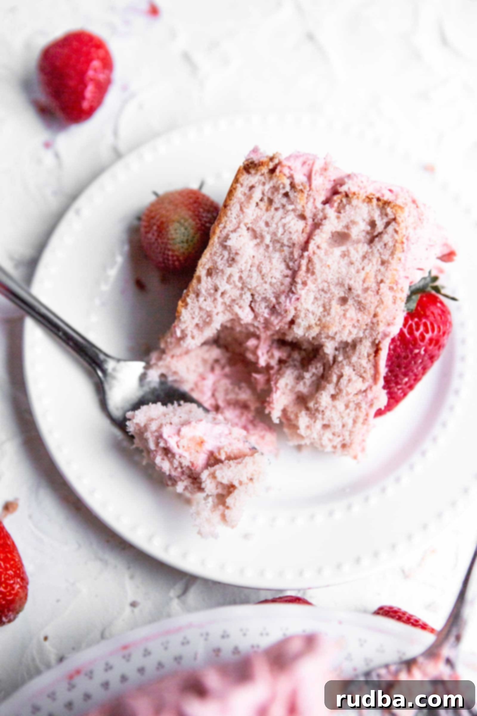 a slice of a two layer pink cake on a white plate with a fork and strawberries on the side.