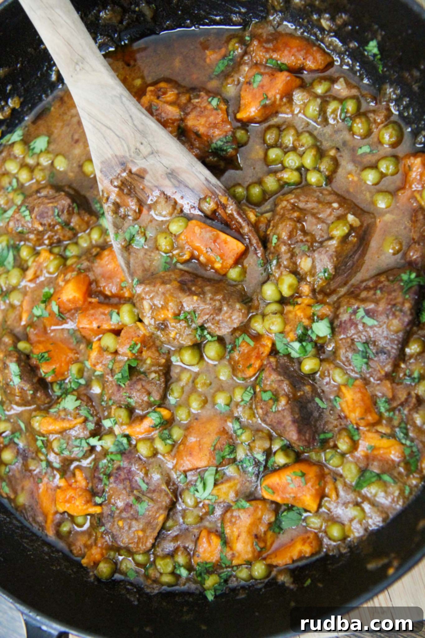 A cast iron skillet filled with hearty sweet potato beef stew, featuring succulent beef cubes, vibrant green peas, and tender sweet potato chunks, accompanied by a wooden spoon.