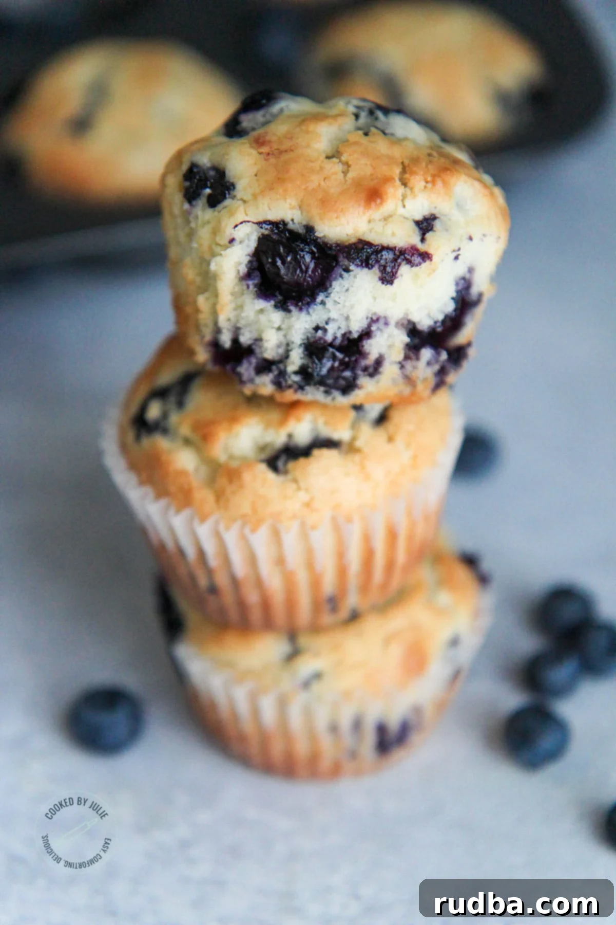 Three perfectly golden brown blueberry muffins stacked, with fresh blueberries scattered around