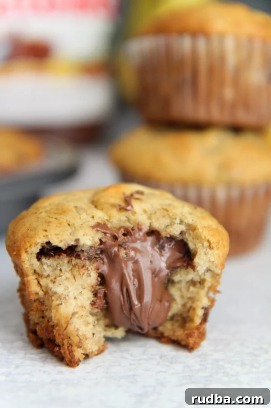 Nutella stuffed banana muffin up close with a nutella jar and more muffins in the background. The muffin is golden brown with a visible Nutella swirl.
