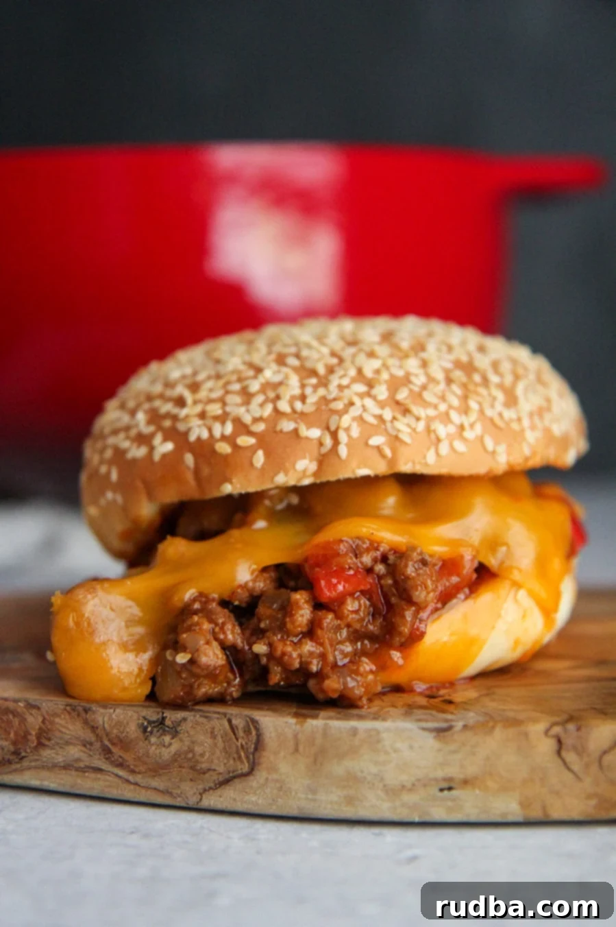 A delicious homemade sloppy joe sandwich with melted cheddar cheese up close, with a vibrant red Dutch oven visible in the softly blurred background.