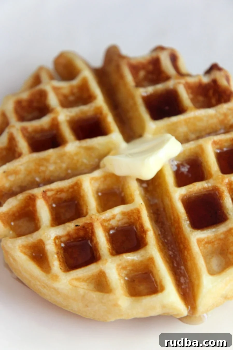 Close-up of a golden-brown buttermilk Belgian waffle on a white plate, generously drizzled with warm maple syrup and topped with a slice of melting butter.