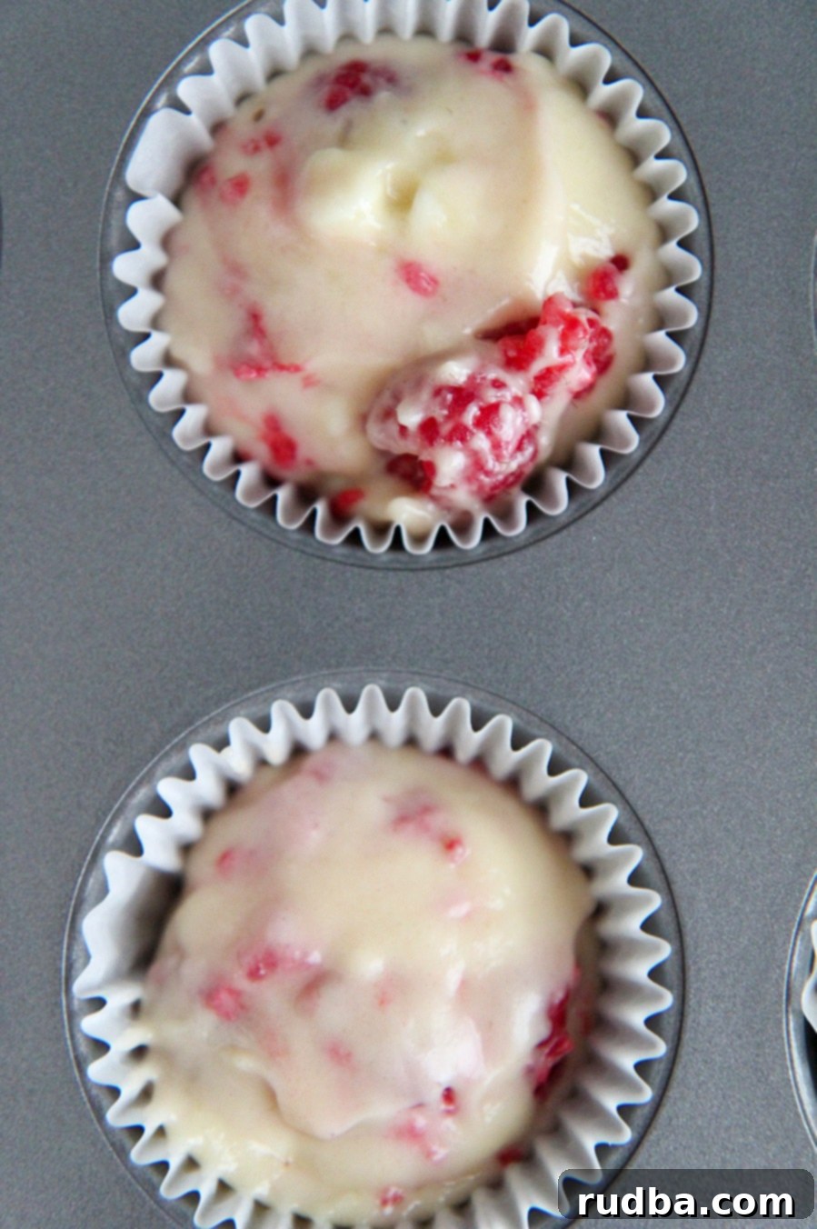 A muffin tin with two cupcake liners filled with white chocolate raspberry muffin batter, ready for baking.