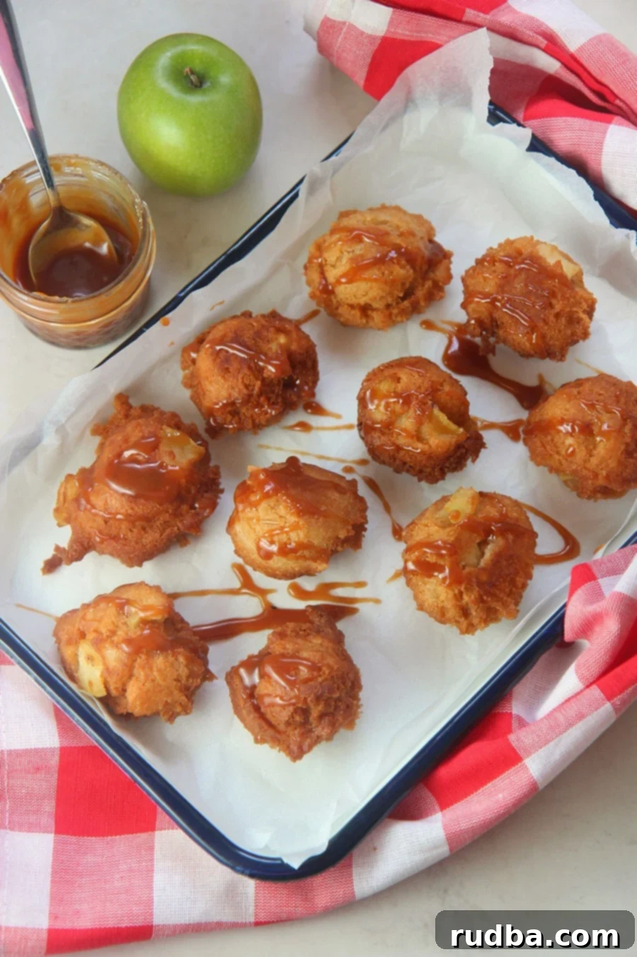 A delicious plate of ten golden-brown apple fritters generously drizzled with rich caramel sauce, accompanied by a small jar of homemade caramel, a fresh green apple, and a rustic red and white towel, evoking a cozy, autumn feel.