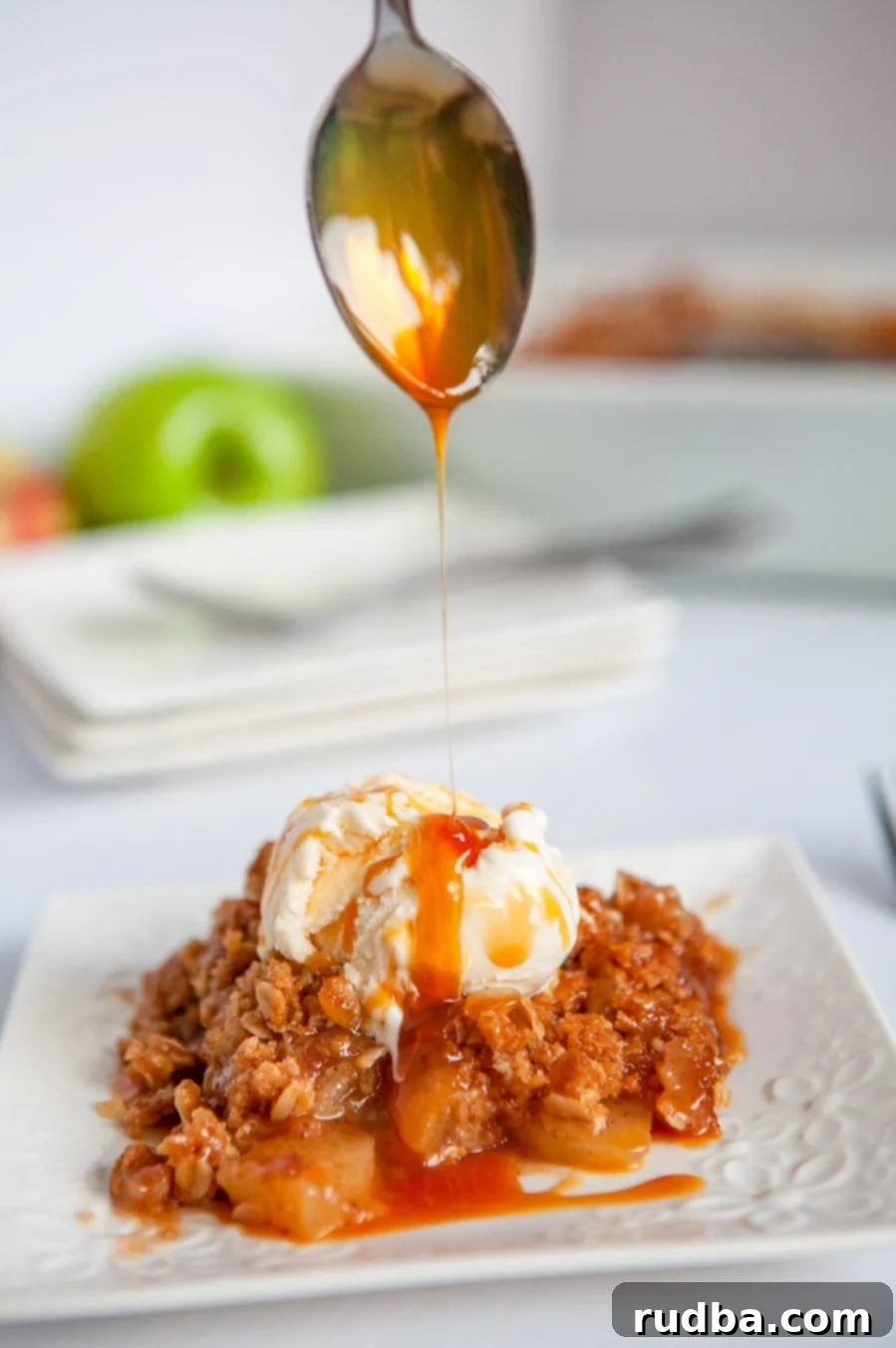 A spoonful of caramel sauce being drizzled over a warm apple crisp, served with vanilla ice cream on a white plate, highlighting the golden crumb topping and soft apples.