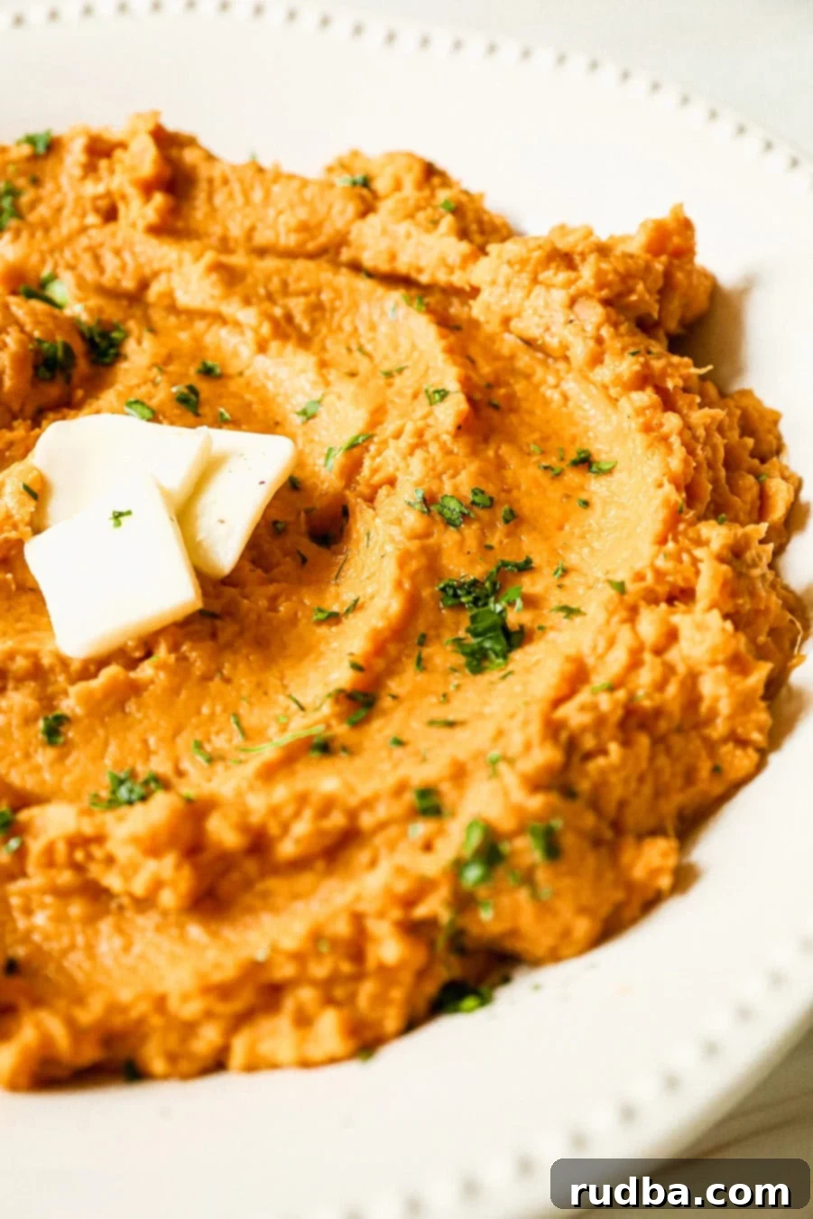 Creamy mashed sweet potatoes served in a white bowl, topped with a pat of melted butter and fresh green herbs like chopped parsley or chives.
