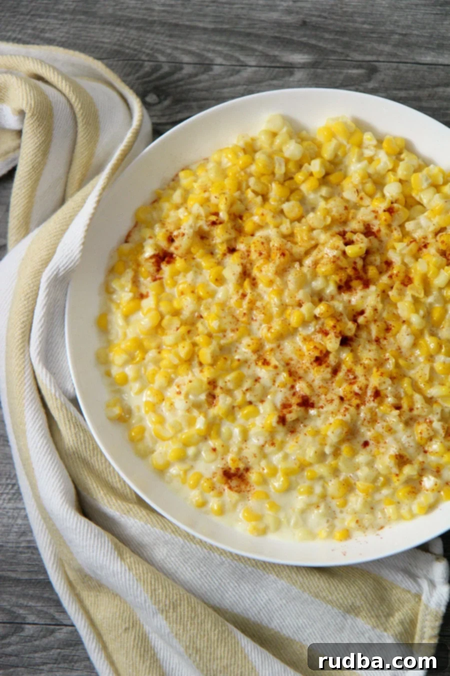 A bowl of creamy homemade creamed corn garnished with fresh parsley, sitting on a white plate with a rustic yellow and white kitchen towel on the side, ready to be served.