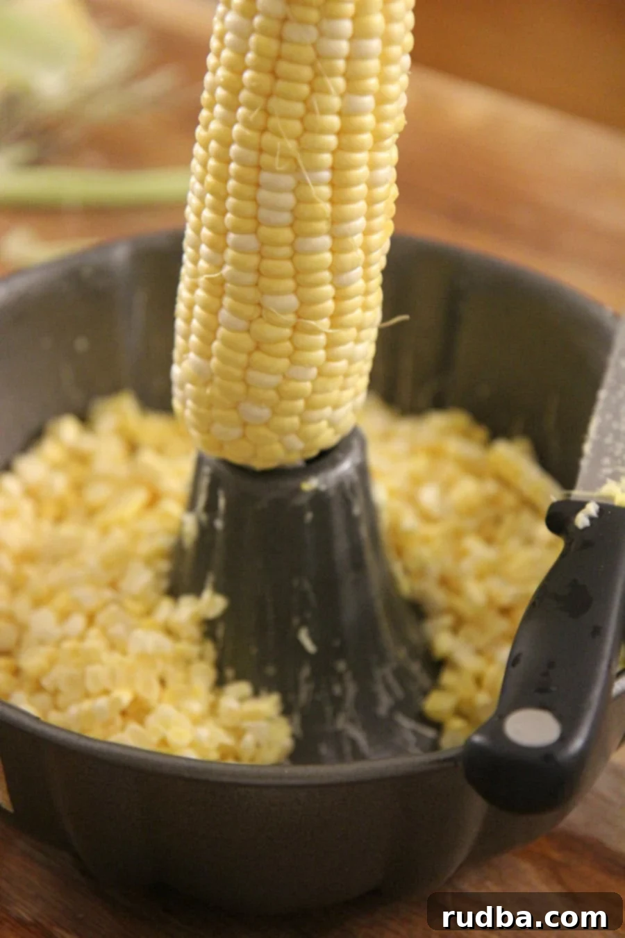 Fresh corn on the cob being prepared, with one ear partially cut to remove kernels, resting inside a bundt pan next to a knife, illustrating the preparation process.
