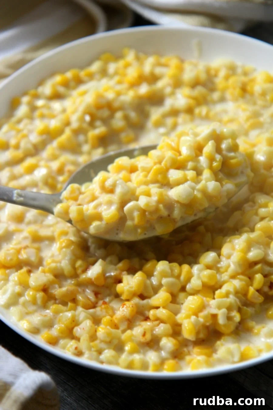 A serving spoon lifts a generous portion of thick, creamy homemade creamed corn from a white bowl, showcasing its rich, velvety texture and golden color.