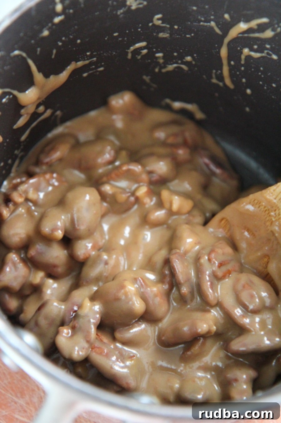 Pecan praline mixture simmering in a black saucepan, with a wooden spoon stirring the rich, caramelizing liquid and nuts.