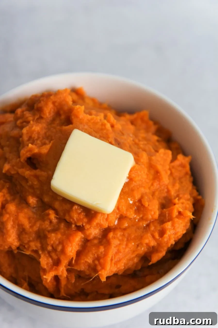 A small white bowl filled with creamy mashed sweet potatoes, topped with a slice of melting butter, ready to be served.
